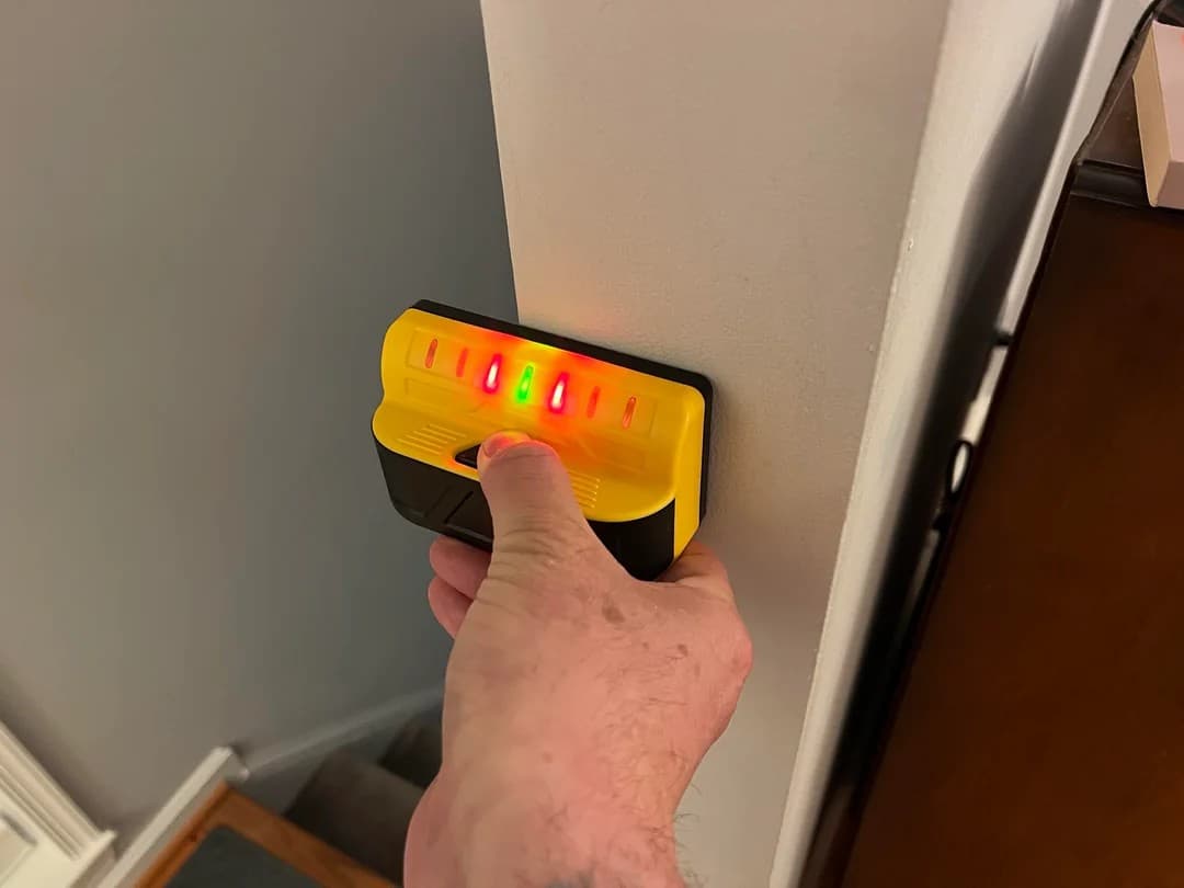 A real wall-check setup with a stud finder, pencil mark, tape measure, outlet reference, and planned drilling area.